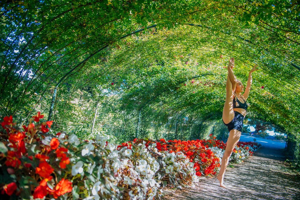 Contortionist performing a leg stretch in a leafy garden tunnel lined with red and white flowers, Tacoma, WA, USA.
