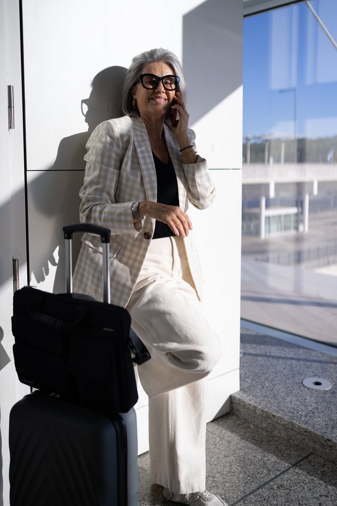 Businesswoman traveler stands inside a modern airport terminal talking on her phone beside a black rolling suitcase in sunlight, Spain, Madrid.