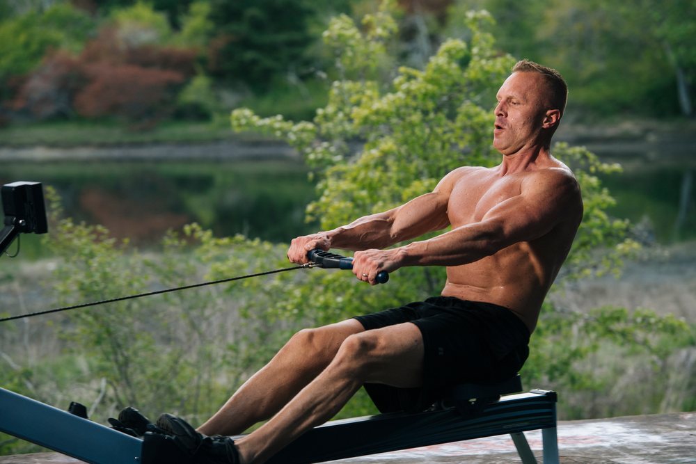 Muscular shirtless man rowing on an exercise machine outdoors near a lake, USA.