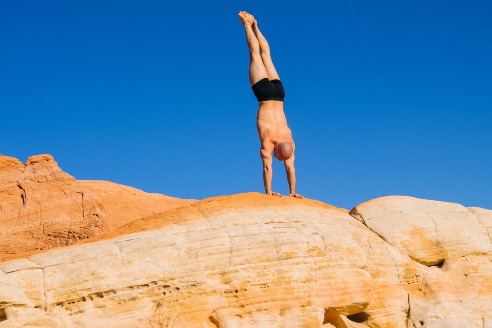 Muscular man with bare torso performs a handstand on a rock formation under a clear blue sky in a USA desert landscape, yoga, acrobatics.
