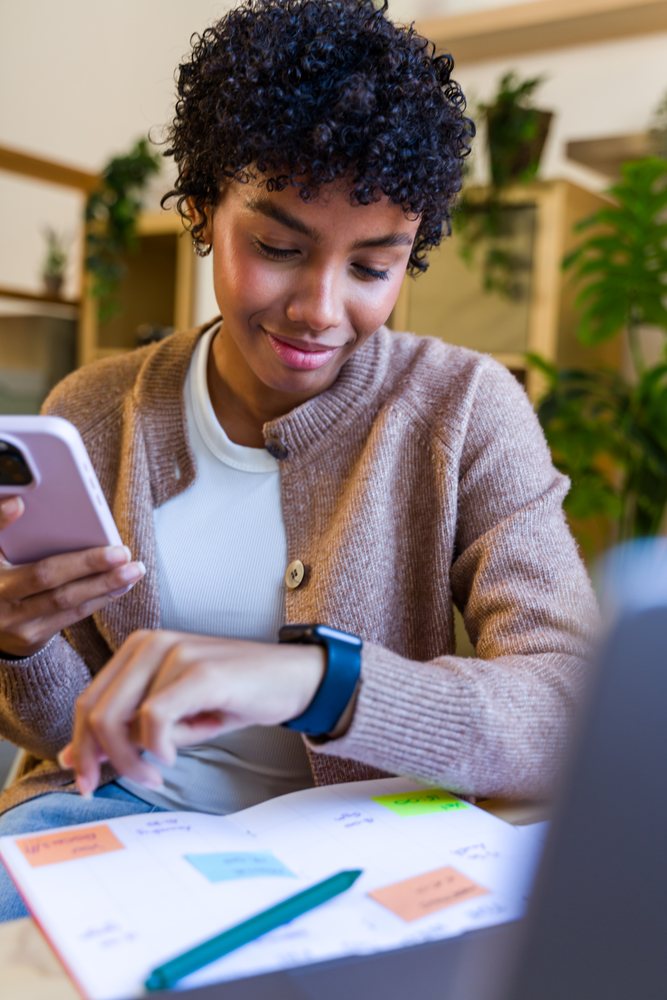 Woman indoors plans at her desk using a smartphone, checking her smartwatch, and writing in a notebook with colorful sticky notes, Spain.