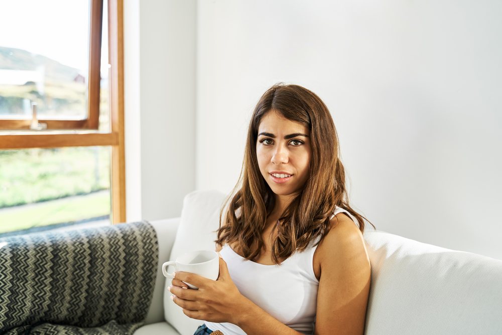 A white woman sitting on a sofa holding a cup in a bright room
