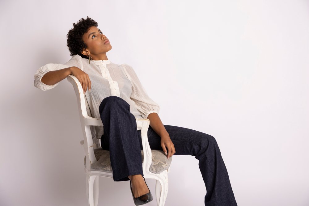 Woman in a white blouse and dark pants sits on a chair looking upwards in a studio setting, fashion, young culture.