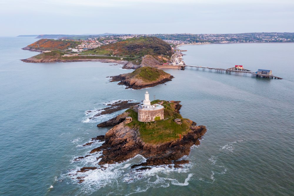 Mumbles Lighthouse stands on a small rocky island at Sunrise near a pier with a ferris wheel and grassy hill by the coastal landscape, Swansea, Glamorgan, Wales.