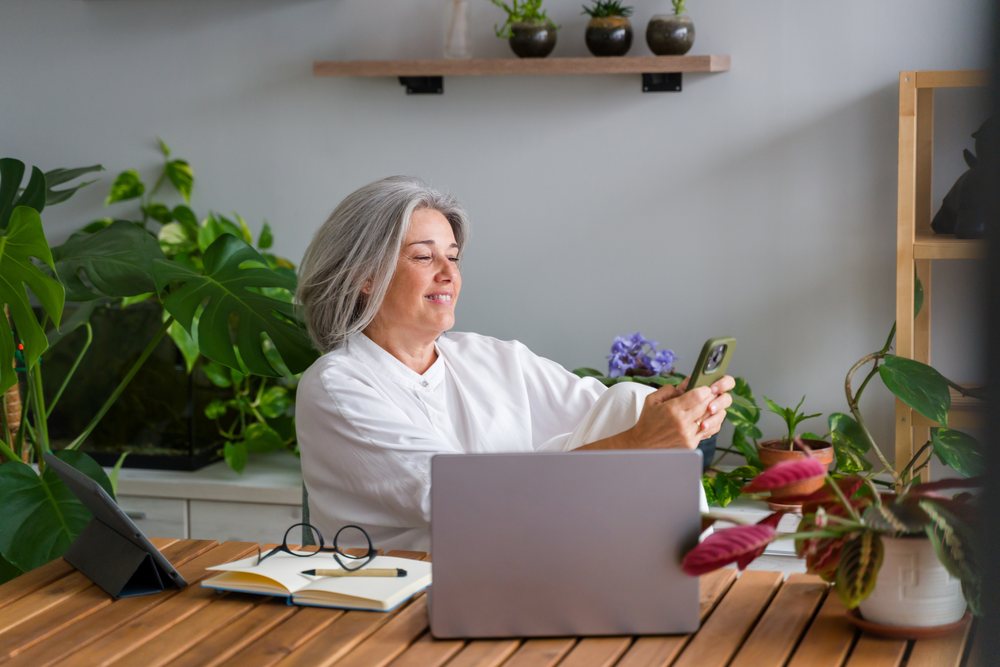 Middle aged woman using smartphone and smiling while sitting at home office desk surrounded by indoor plants, Spain.