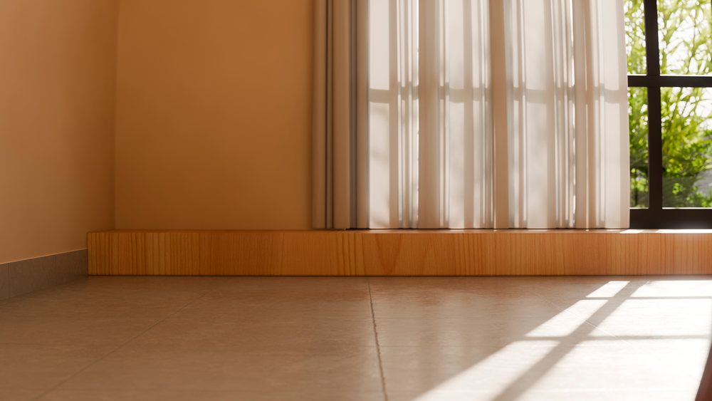 Sunlit empty room with tiled floor, beige walls, and sheer curtains over a window revealing green plants outside.