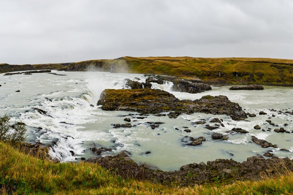 A panoramic view of a rocky river with cascading waterfalls and yellow grassy hills under an overcast sky, Iceland.