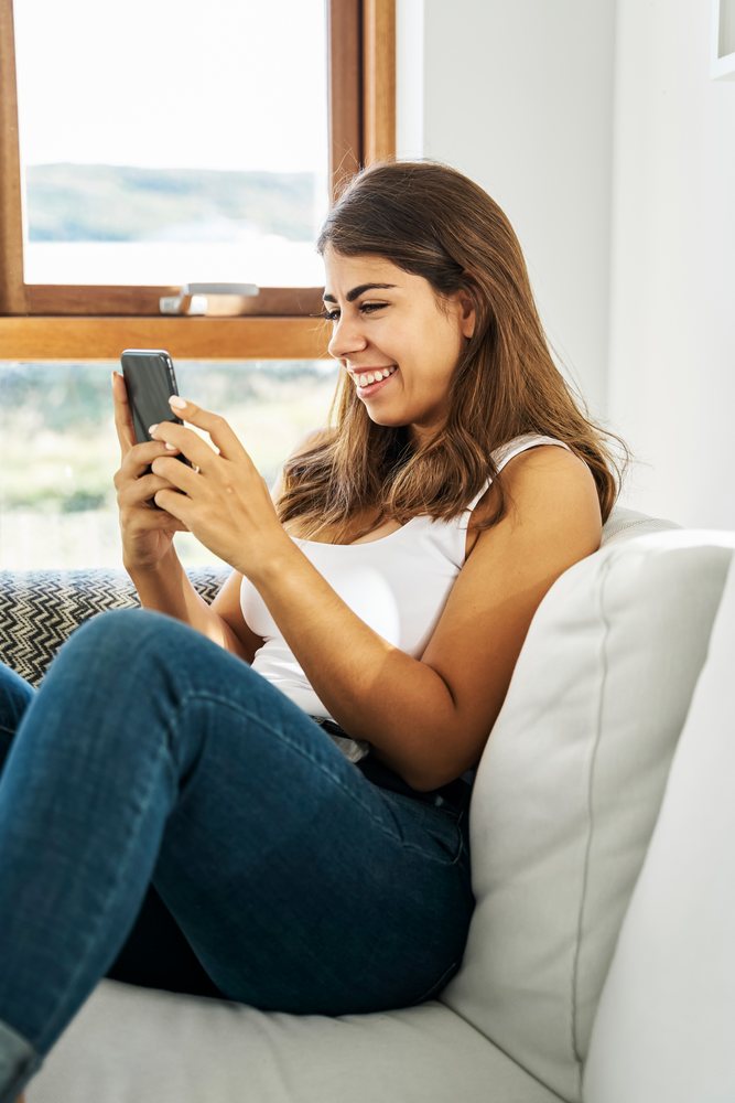 A white woman smiling while using her mobile phone on a couch