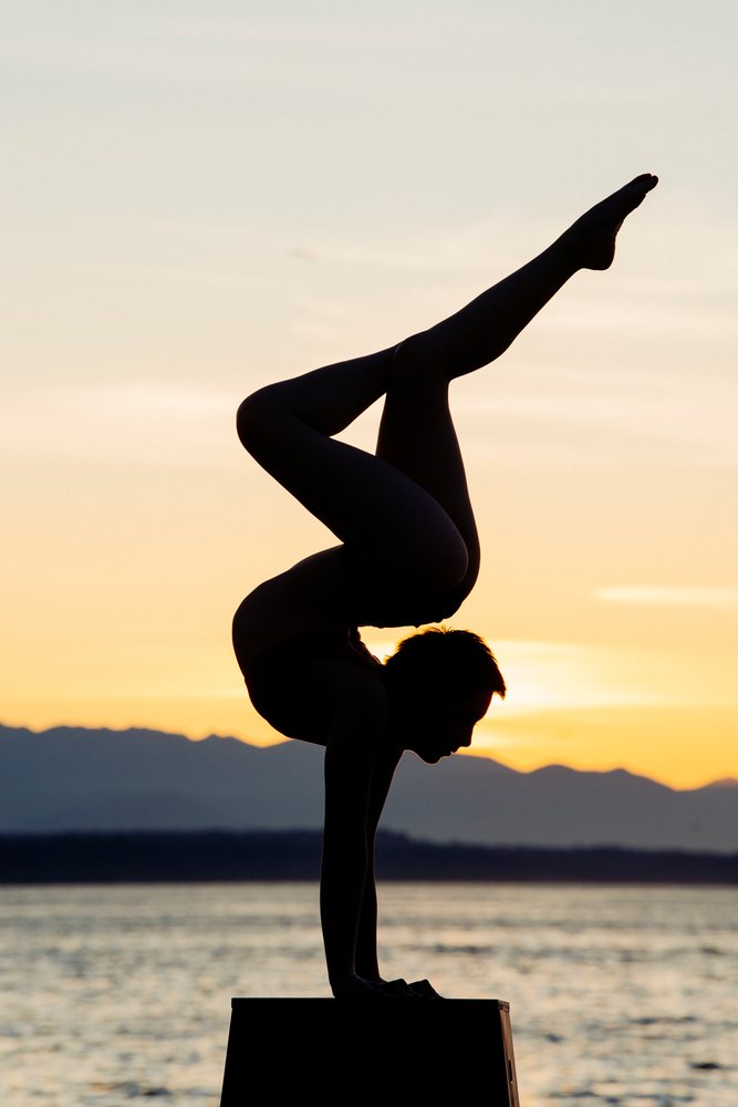 Person performing a handstand yoga pose silhouetted against a sunset over water with mountains in the background, USA.