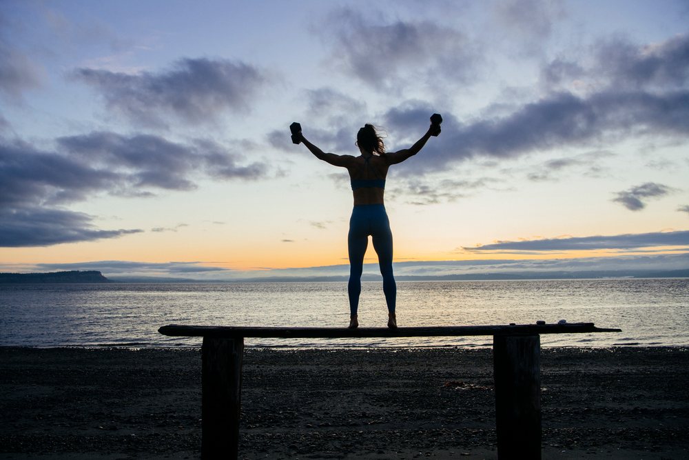 Silhouette of a woman exercising with dumbbells on a balance beam at sunset on a calm beach, WA, USA.