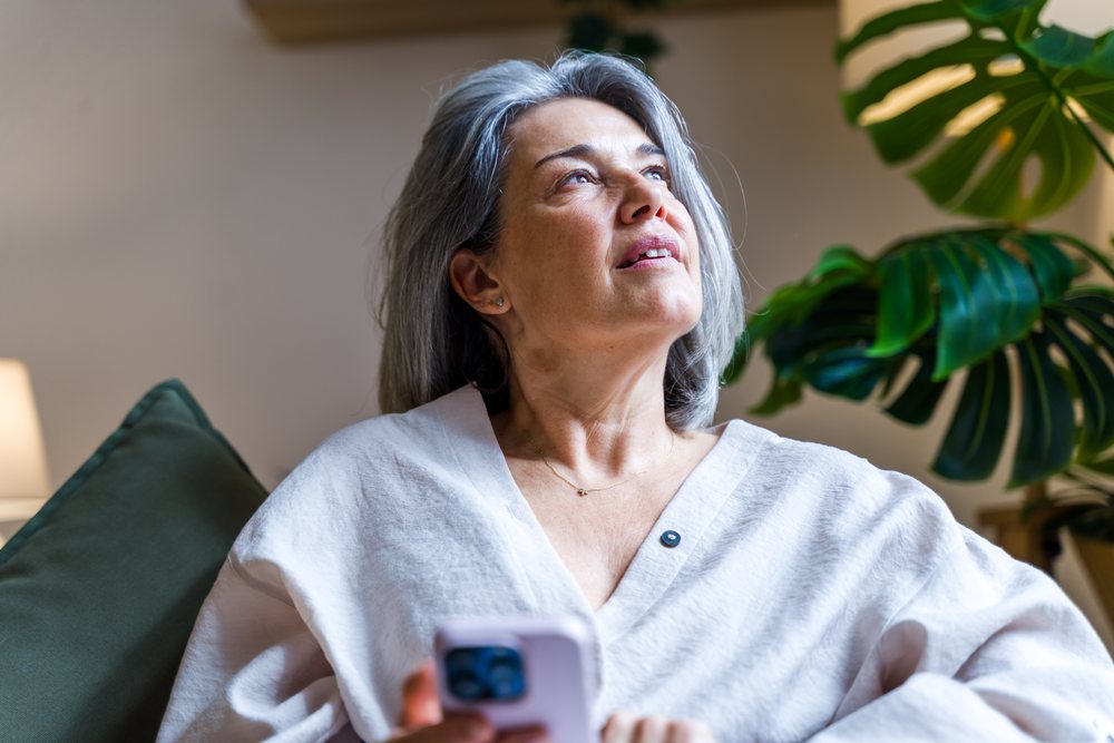 Middle-aged woman with gray hair sits indoors holding a phone and looking up thoughtfully beside a green pillow and indoor plant, Spain.