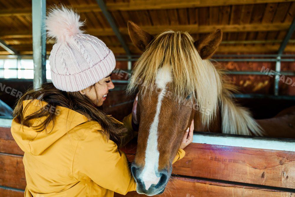 A white woman gently pets a brown horse inside a wooden stable