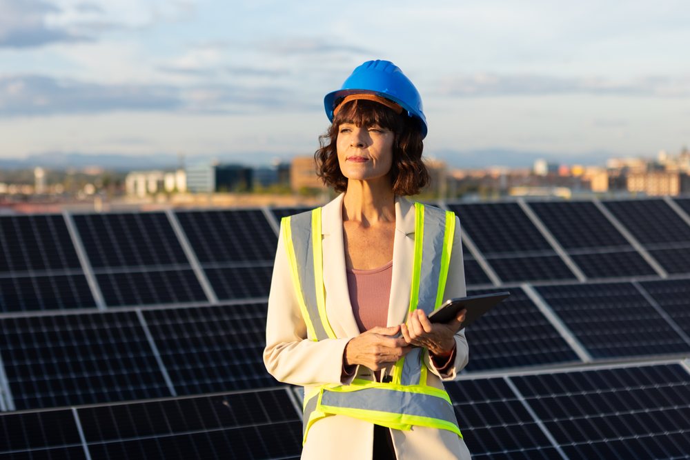 Woman engineer inspects solar panels outdoors at a solar farm, holding a tablet and wearing a blue hard hat and reflective safety vest, Madrid, Spain.