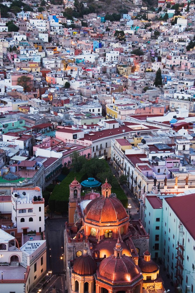 Dusk cityscape of Guanajuato, Mexico showing colorful buildings, historic church domes, and a street with Hotel San Diego sign visible.