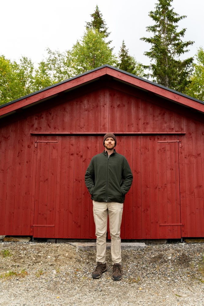Man wearing a green jacket and brown hat stands outside a red wooden shed, Norway.
