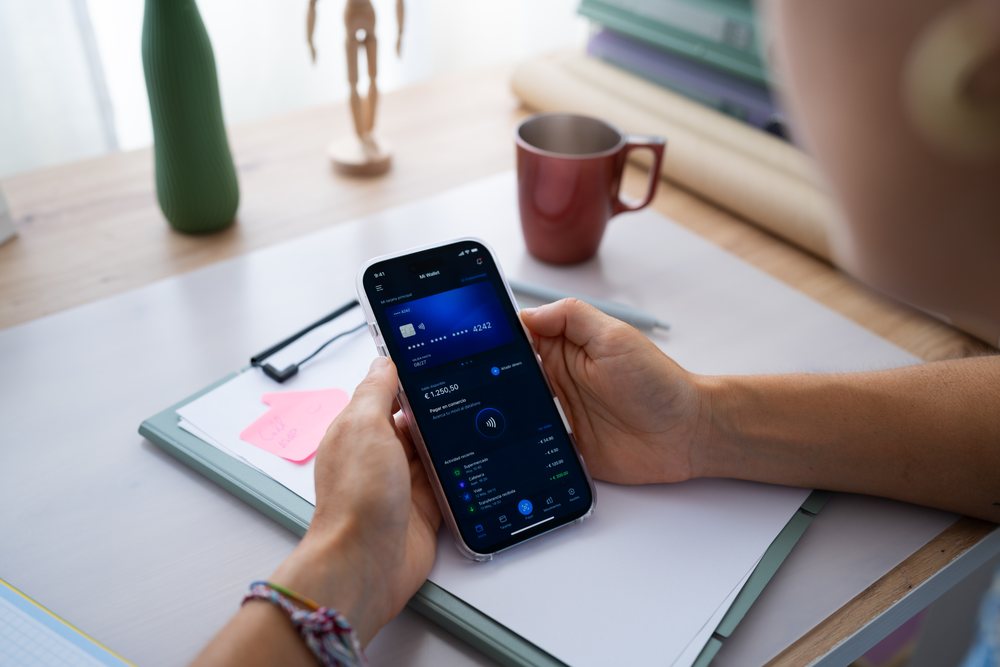 Person holding a mobile phone displaying a digital wallet app on a desk with office supplies and a red mug, telecommuting, Spain.