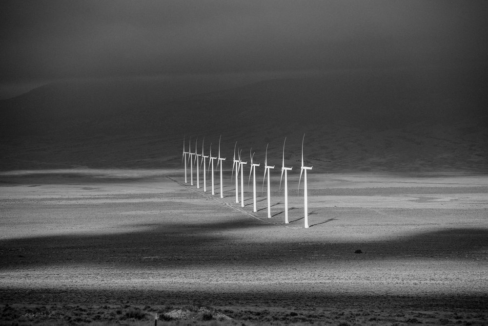 Line of wind turbines stretches across a Nevada desert landscape with mountains and a cloudy sky casting shadows, USA.