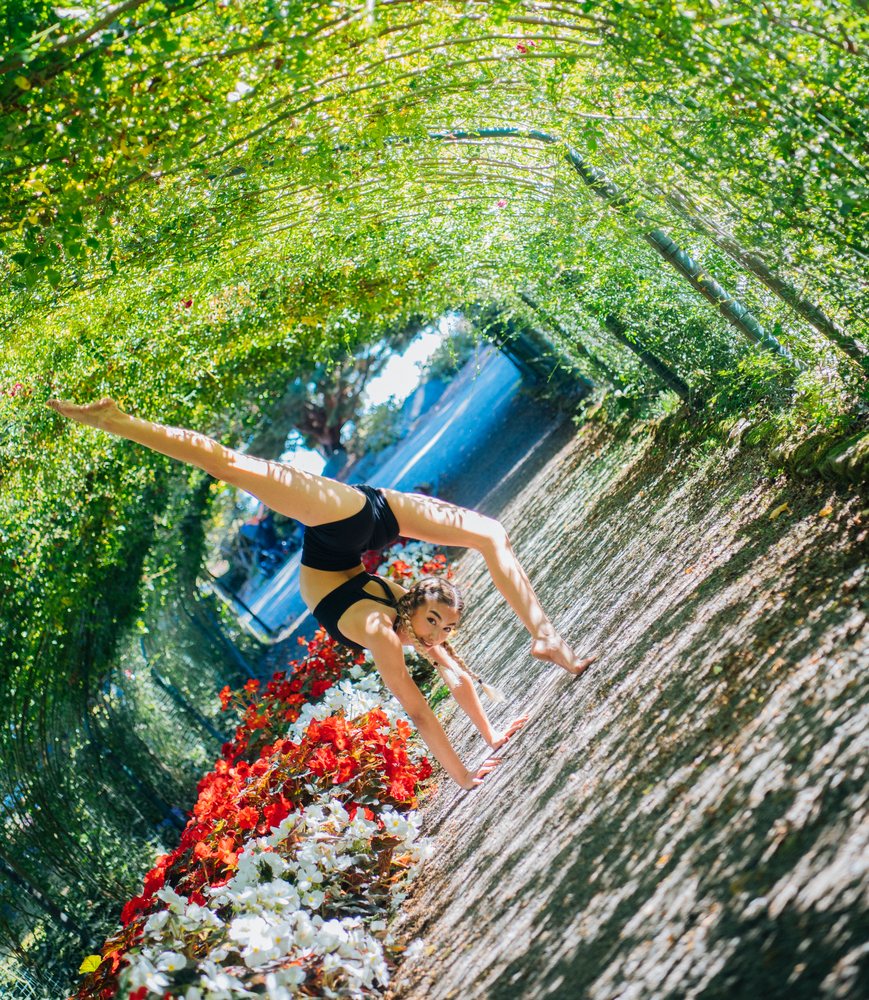 Woman performing a contortion handstand under an arched garden pathway beside a flower bed with red and white flowers, contortionist, Tacoma, WA, USA.