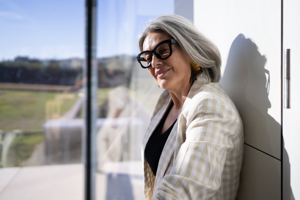 Mature woman with gray hair and glasses smiling and resting indoors by a window in natural daylight, businesswoman, traveler, Spain, Madrid.