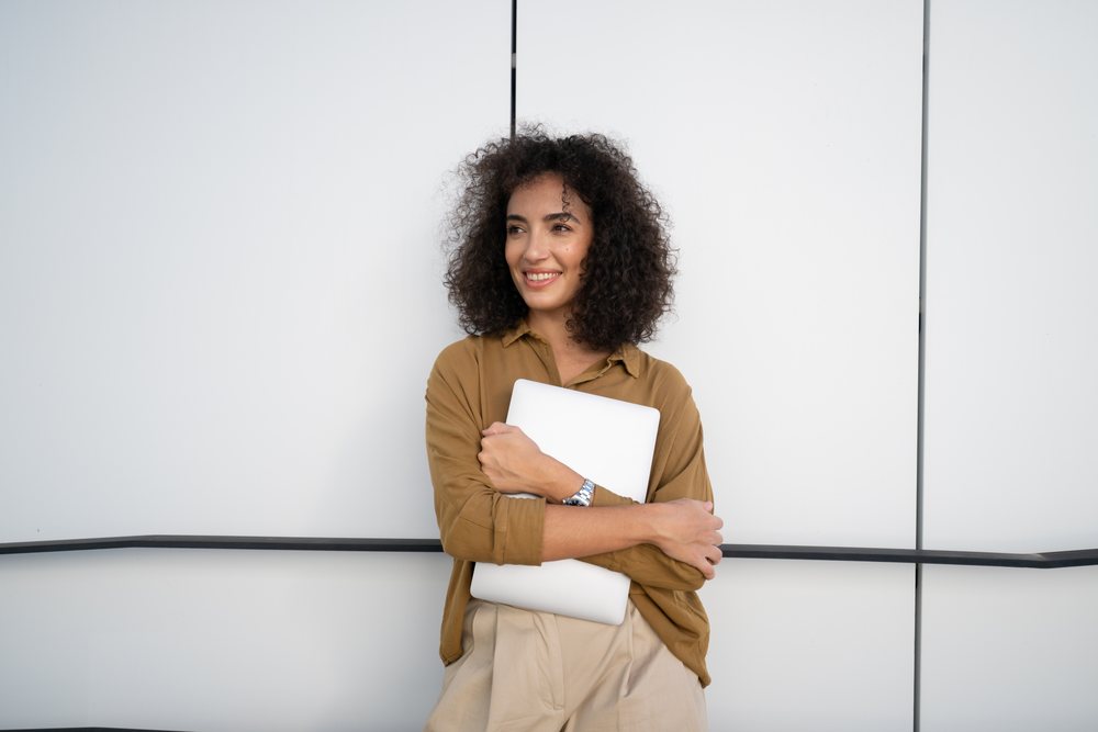 Woman with curly hair wearing a brown shirt and beige pants smiles while holding a laptop computer outdoors against a white wall, Madrid, Spain.