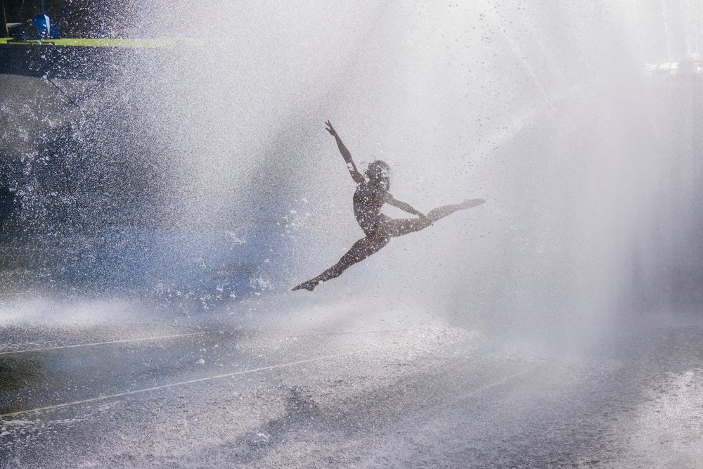 Dancer performing a high leap surrounded by water spray outdoors, partner, acrobatics, yoga, USA.