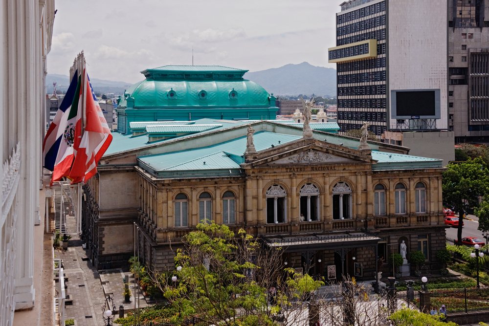 Ornate National Theatre historic building with green roof and rooftop statues, with urban cityscape and mountains behind, San Jose, Costa Rica.