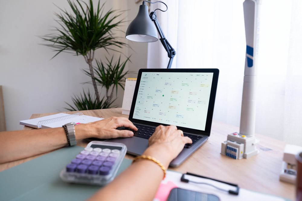 Person typing on a laptop with a calendar app open at a wooden desk in a home office setting with natural light, Spain.