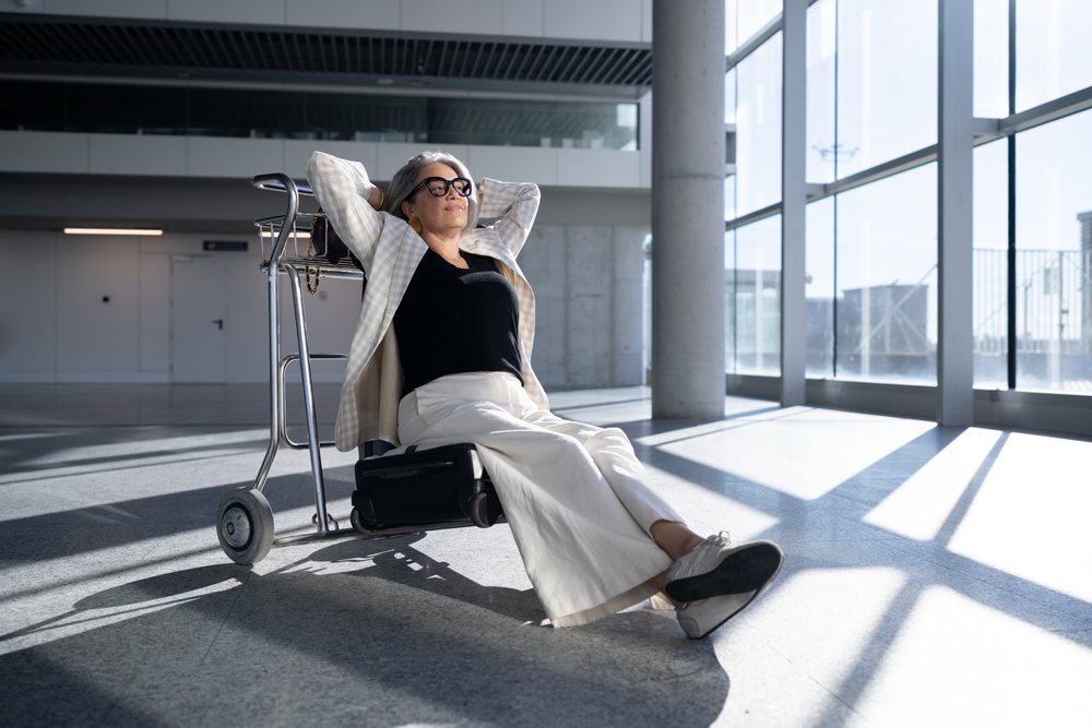 Mature woman with gray hair and glasses relaxes on a cart inside a modern airport terminal, with sunlight streaming through large glass windows, businesswoman, traveler, Spain, Madrid.