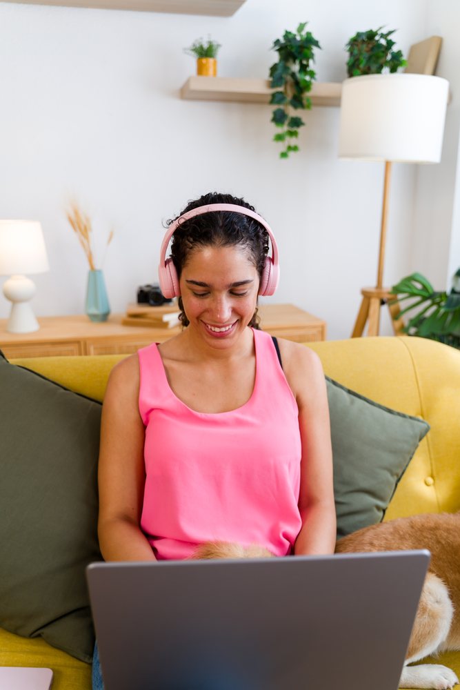 A woman wearing pink headphones and a pink top smiles while using a laptop on a yellow sofa with a dog lying beside her in a living room, Spain.