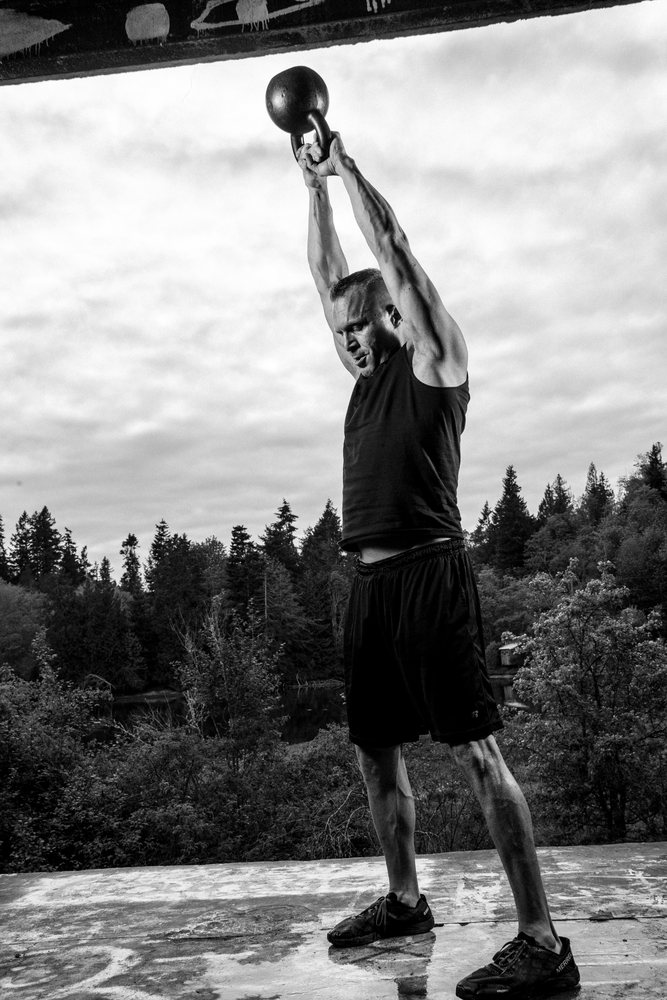 Man lifting a kettlebell overhead during an outdoor strength workout in a forested natural setting, USA.