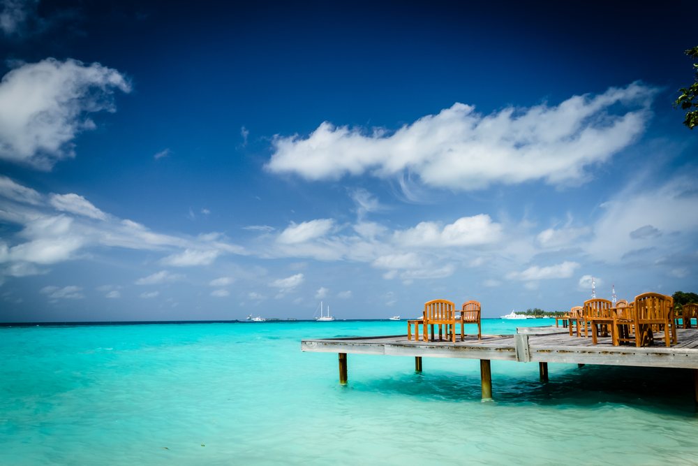 Wooden dock with wooden chairs extending over turquoise ocean water under a blue sky with white clouds, summer, Holliday, travel, landscape, Maldives.