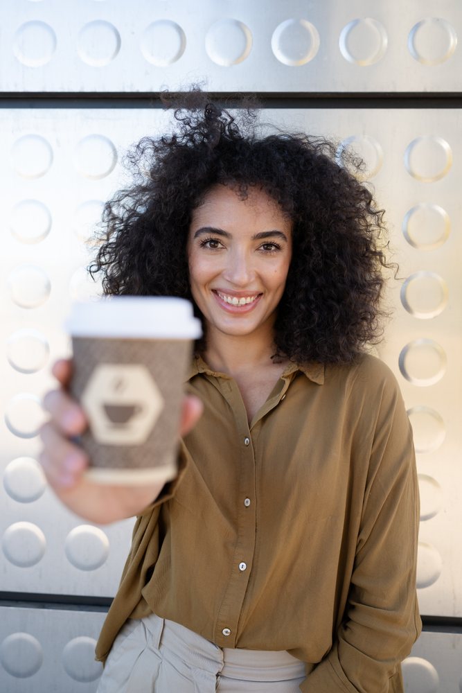 Woman with curly dark hair smiles while holding a takeaway coffee cup outdoors, Madrid, Spain.