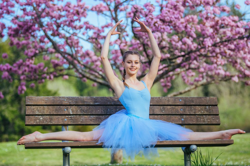 Ballet dancer in a blue leotard performs a split while sitting on a bench in a spring park with pink blossoming trees, USA.