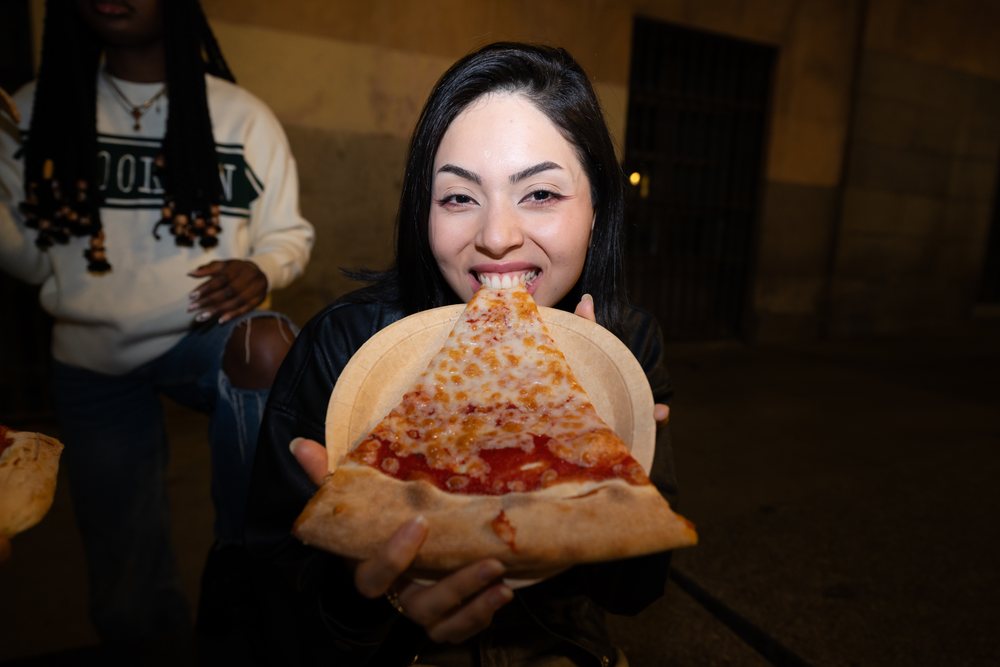 Woman eating a large slice of cheese pizza topped with tomato sauce outdoors at night on an urban street, Madrid, Spain.