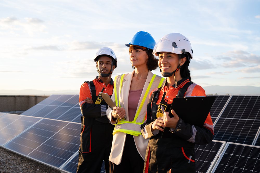 Three engineers including a woman manager in a reflective vest stand by solar panels outdoors at sunset, Madrid, Spain.