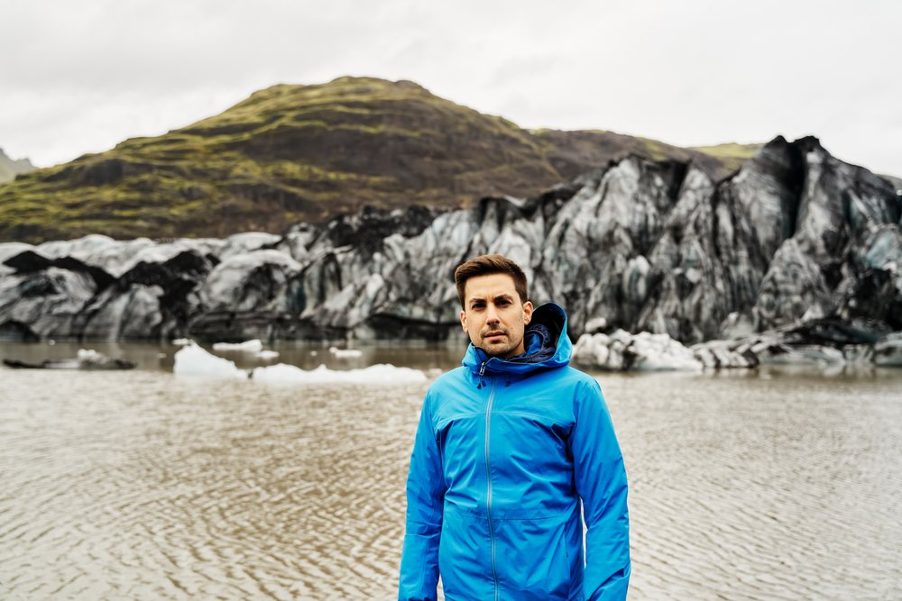 A white man in a blue jacket stands by a glacier lake and mountains