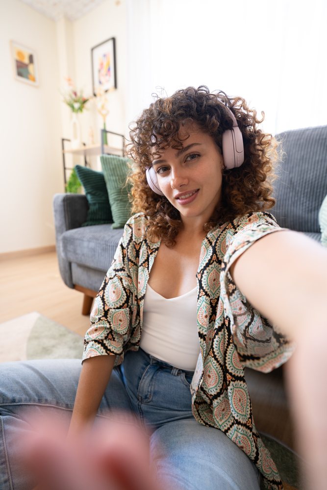 Young woman with curly hair wearing wireless headphones takes a selfie while seated on a sofa in a cozy living room with flowers and wall art, home, girl, Spain.