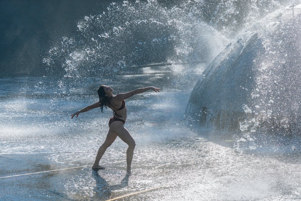 Woman in swimsuit dancing joyfully in a water fountain with sunlight highlighting splashing water, partner, acrobatics, yoga, USA.