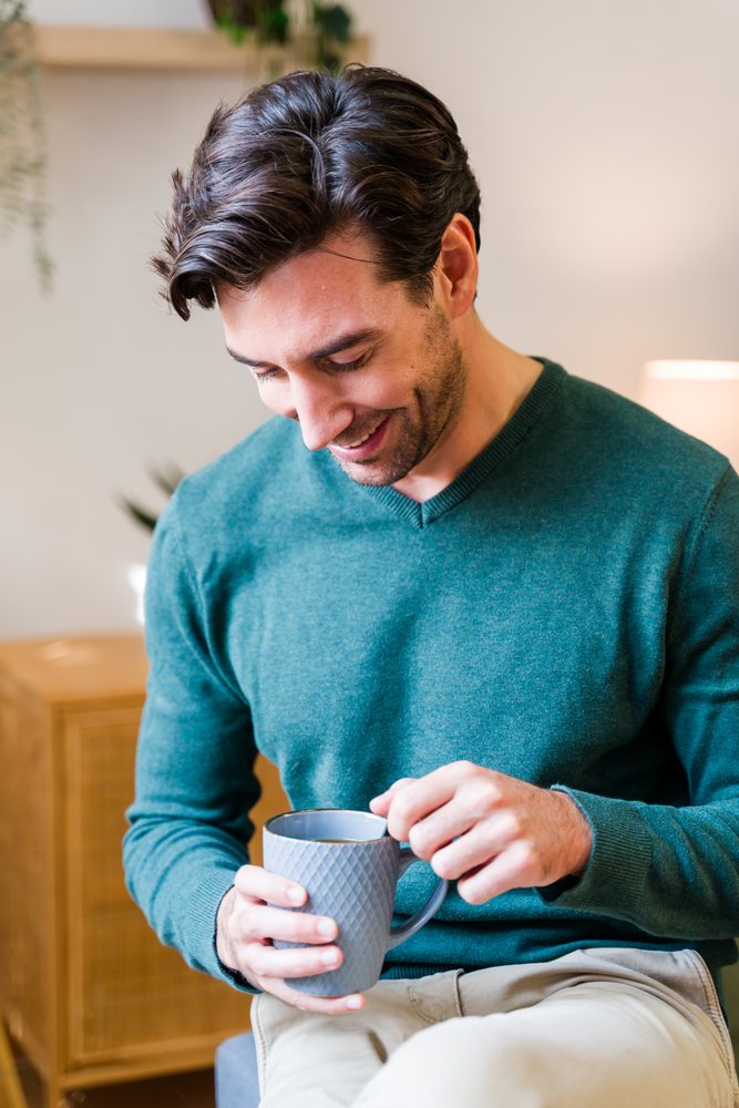 Man in a green sweater smiling indoors while holding a gray mug near a wooden cabinet with a lamp and plant, Spain.
