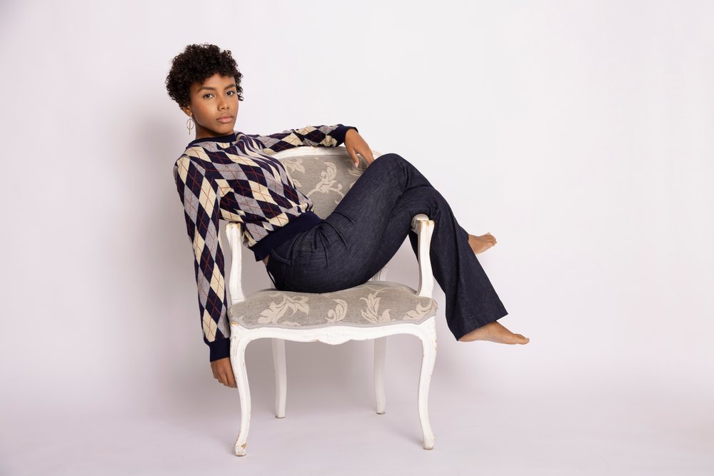 Young woman wearing a blue, gray, and beige argyle sweater and dark blue jeans seated barefoot on a floral-upholstered white ornate armchair in a studio with white background, fashion, young culture.