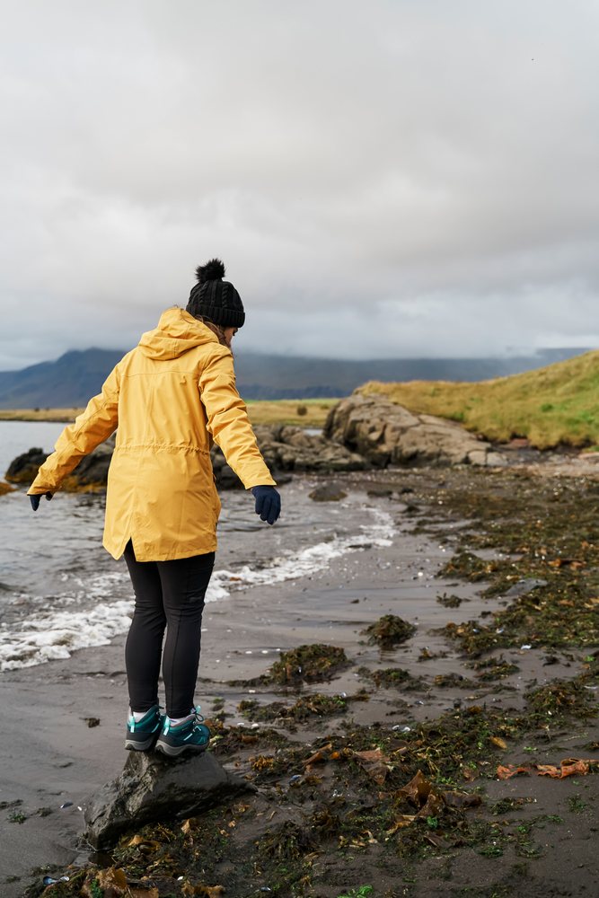 A white woman balances on a rock along the shoreline