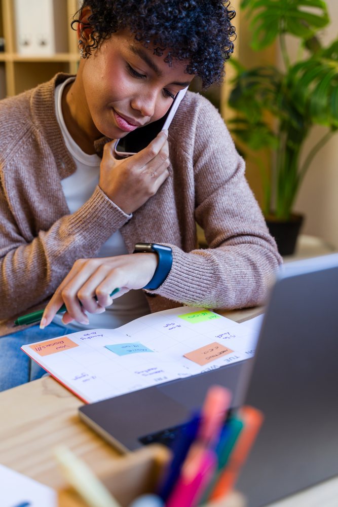 Woman making a phone call while checking a calendar and writing at her desk in a home office, Spain.