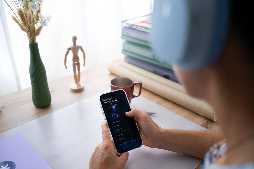 Person wearing headphones using smartphone to play music at a desk decorated with a wooden mannequin, green vase with white flowers, and coffee cup, telecommuting, Spain.