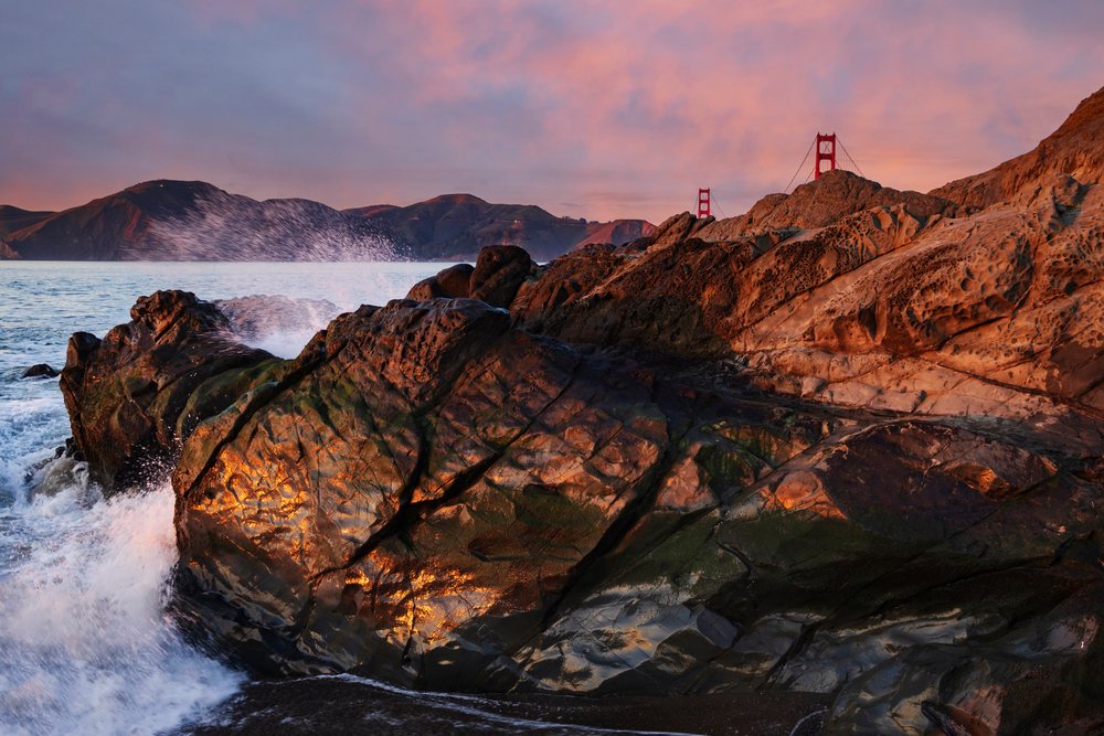 Rocks along the ocean shore at Baker Beach with waves splashing and the Golden Gate Bridge visible at Sunset, landscape, cityscape, city life, San Francisco, California, USA.
