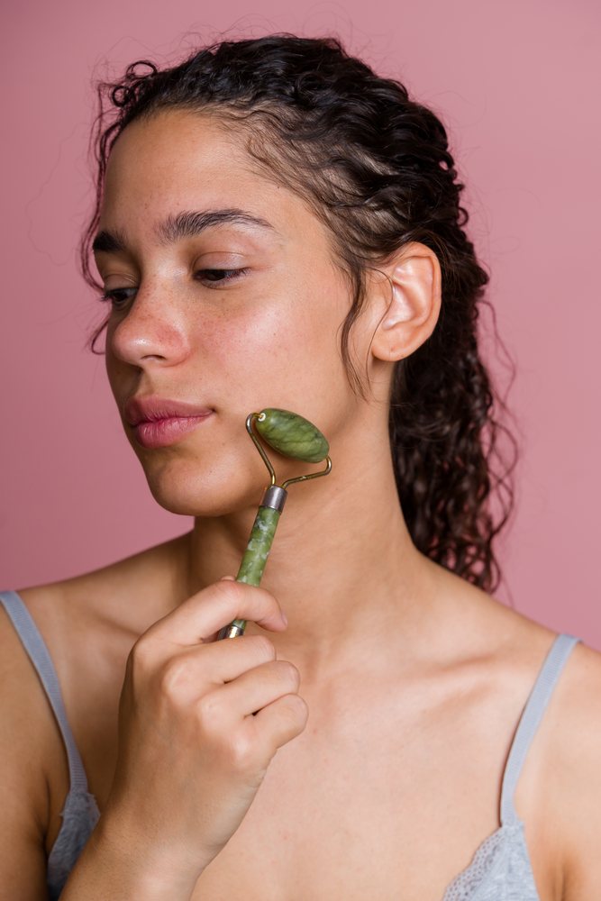 Woman with curly hair using a jade roller for facial massage against a pink background during a beauty shoot, Spain.