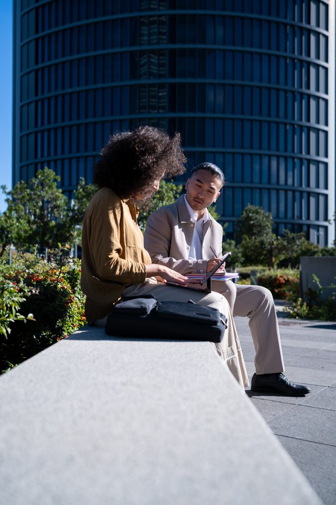 Two men seated on a bench in an urban park using a tablet and writing in a notebook with a modern building in the background in sunlight, Madrid, Spain.