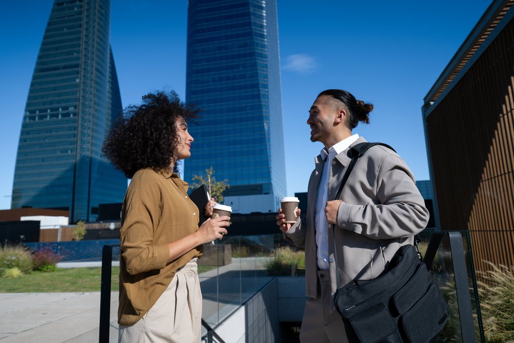 Two people, a man and a woman, holding coffee cups and talking outdoors by glass railings with office towers, Madrid, Spain.