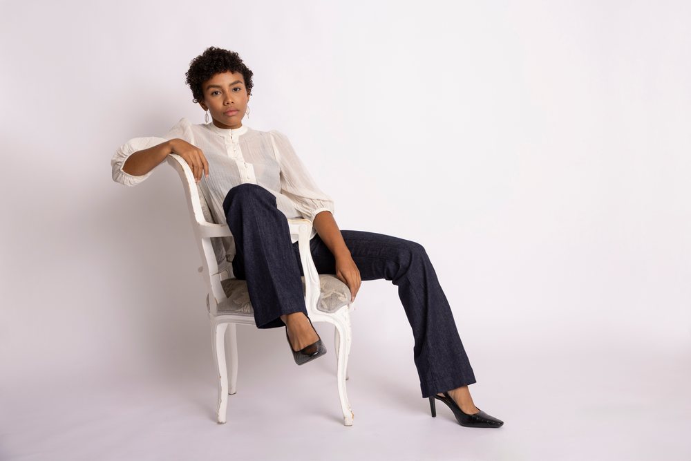 Woman wearing a white blouse, dark jeans, and black high heels sits on a white cushioned chair in a plain white studio, fashion, young culture.