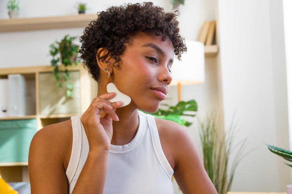 Young woman performing facial massage with a cupping tool indoors at home with plants in background.