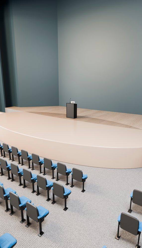 Empty indoor lecture hall featuring a conference stage with podium and microphone, rows of blue seats, and beige carpet flooring.